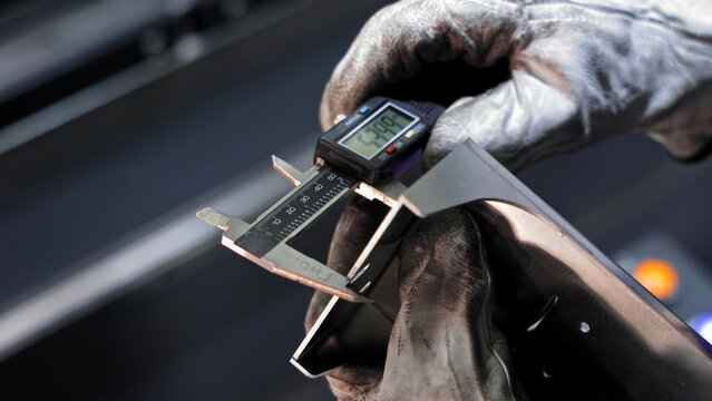 Precision measurement of metal part with digital caliper. Worker wearing gloves using a digital caliper to accurately measure the dimensions of a machined metal component in a workshop.