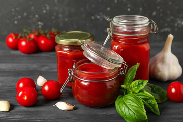 Jars of tasty ketchup with garlic, tomatoes and basil leaves on wooden table against black grunge wall