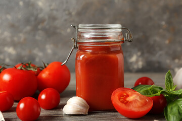 Jar of tasty ketchup with garlic, fresh tomatoes and basil leaves on wooden table against grunge grey wall