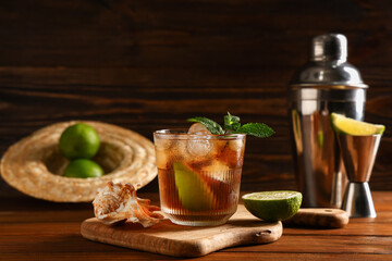 Glass of cold Cuba Libre cocktail with seashell, measure cup and shaker on wooden background