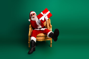 Santa Claus sitting on a golden chair holding a red gift, wearing sunglasses, in front of a vibrant...