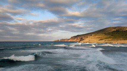 Obraz premium Horizontal photo of stormy sea waves rolling into a bay surrounded by green mountains under a cloudy sky. Dramatic ocean landscape with moody weather and powerful nature energy. Perfect for travel