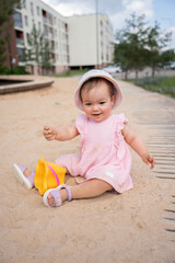 cheerful toddler in a pink dress and white hat playing in urban playground's sandy area with colorful toys on a sunny day, surrounded by residential buildings and greenery, curiosity, family