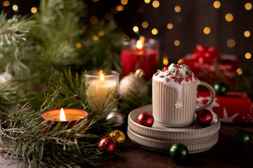 cozy christmas scene with festive decor featuring a mug of hot chocolate topped with whipped cream and sprinkles, surrounded by pine branches, candles, ornaments, and glowing holiday lights, season