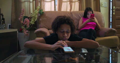 Young boy leaning on glass table staring at smartphone, expressionless, teenage girl behind immersed in her phone, emotional detachment and social media addiction depicted
