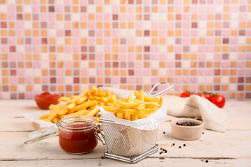Basket for deep fryer with tasty french fries and jar of ketchup on light wooden table against tile wall