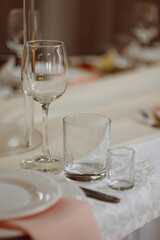 Three empty glasses of different shapes on a beige tablecloth in a softly lit dining setting with white chairs. Concept hospitality, event catering, table setting, restaurant service.