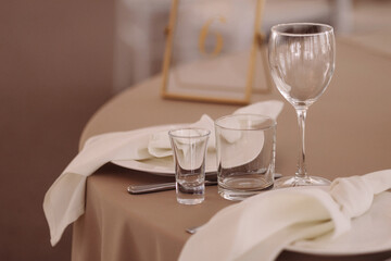 Three empty glasses of different shapes on a beige tablecloth in a softly lit dining setting with white chairs. Concept hospitality, event catering, table setting, restaurant service.