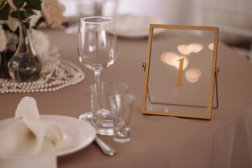 Close-up of elegant wedding table setup with glasses, white napkin, flowers and gold frame table number. Warm light and beige tablecloth create festive mood. Concept of celebration, decor, reception.