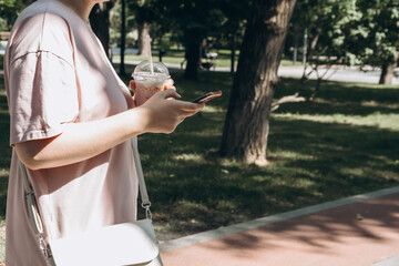 Person outdoors holding a smartphone and a cold drink, wearing casual attire. The park setting suggests a tranquil atmosphere, with trees and a pathway in the background.