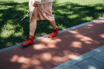 Woman in a gingham skirt and vibrant red shoes walking on a sunny path. Her legs are in motion, casting playful shadows on the sidewalk as greenery lines the path.