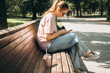 Obraz premium Woman sitting on a wooden bench in a sunny park, focused on her smartphone. She enjoys a refreshing drink by her side, enjoying leisure time outdoors in the shade of trees.