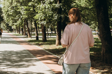 Woman strolls along a tree-lined path in a park on a sunny day. Wearing casual clothes and carrying a crossbody bag, she enjoys the peaceful outdoor atmosphere surrounded by greenery.