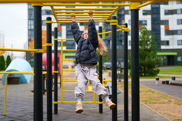 child on monkey bars at playground in urban setting. little girl wearing winter jacket and boots, enjoying active outdoor playtime. bright equipment contrasts with modern buildings
