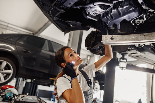 A highly skilled female mechanic thoroughly inspects a vehicles undercarriage using a powerful flashlight