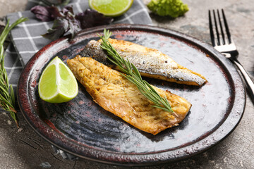 Plate with delicious cooked seabass fish, rosemary and lime slice on grey grunge background