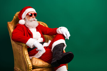 Stylish Santa Claus in a festive Christmas costume relaxing with sunglass on a luxurious chair against a green background