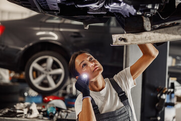 A woman examines a car's underbody with a flashlight in a busy garage, showcasing automotive expertise.