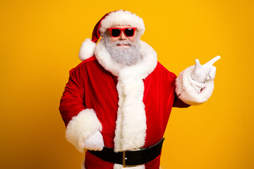 Trendy Santa Claus in red costume wearing sunglass posing against yellow background, making pointing gesture with gloved hand