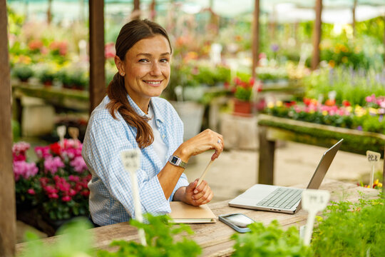 A joyful woman is happily engaging with her laptop while surrounded by vibrant flowers in a lovely garden center - Powered by Adobe