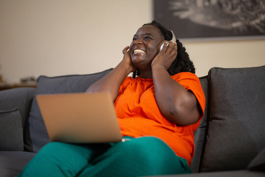 Joyful Black woman with eyes closed enjoying music on headphones while relaxing on the couch with a laptop