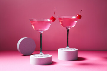 Plaster podiums and glasses of tasty Cosmopolitan cocktail garnished with cherries on pink background