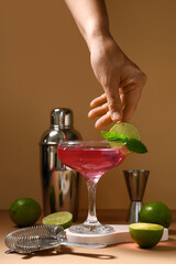 Female hand with glass of tasty Cosmopolitan cocktail, lime and bartender tools on brown background