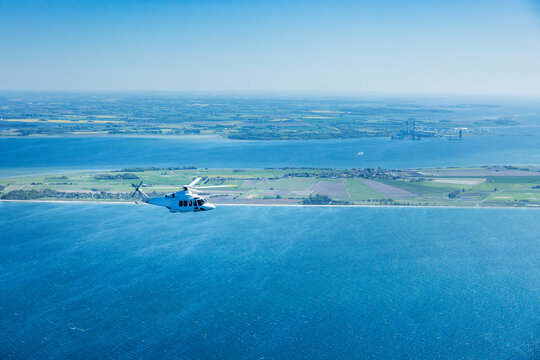 Large helicopter flying over the Baltic Sea with landscape and wind farms in the background.