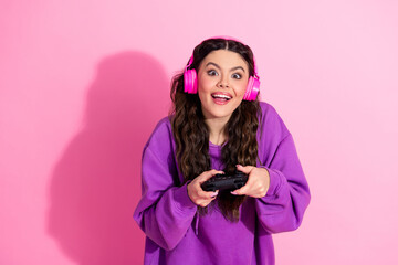 Young woman wearing headphones and playing video games on a pink background, embracing fun and...