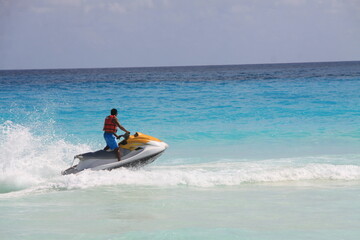 Man riding a yellow and white jet ski across turquoise tropical waters, wearing a red life vest and blue shorts, creating a splash near the beach under a sunny sky.