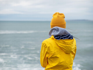 Young teenager tourist in yellow jacket standing by the ocean. Selective focus. Aran islands in the background. Burren area, Ireland. Cloudy sky. Explore Irish nature concept. © mark_gusev