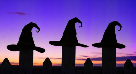 Three silhouettes of witch hats on fence posts against purple sunset sky  
