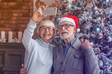 Happy senior couple taking selfie near decorated Christmas tree at living room. Caucasian old elderly couple spouses grandparents vlogging blogging having videocall on New Year Eve