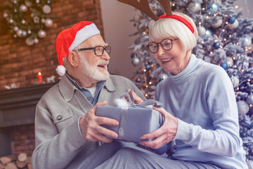 Happy senior couple in Santa hats exchanging Christmas gifts at home. Elderly old grandparents spouses grandfather and grandmother sharing giving presents