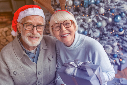 Senior couple with present box sitting in front of Christmas tree and looking in camera. Caucasian old elderly grandparents spouses celebrating New Year together at home