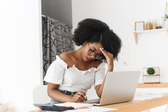 African American woman sitting at home table with laptop, papers, and calculator, looking stressed and upset. Concept of financial problems, remote work challenges, budgeting, and personal finance - Powered by Adobe