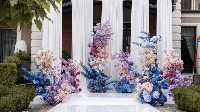 wedding romantic flower arch on a white podium outdoors with fabric in blue pink purple white colors. Delphinium, rose, hydrangea, chrysanthemum, carnation, dahlia