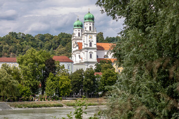 A summer view of St. Stephen&rsquo;s Cathedral in Passau, Germany, seen from the Innstadt side across the river Inn, surrounded by green trees and historic architecture under a cloudy sky.