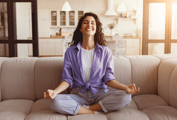 A young woman practices meditation on a comfortable sofa in a bright, modern living room. She has a peaceful expression, enjoying the calming atmosphere created by the sunlight.
