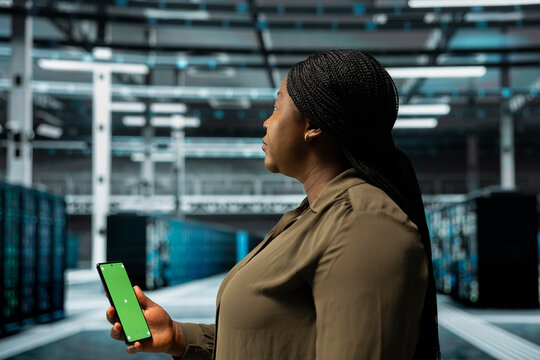 Woman in data center checking mainframes performance data on mockup smartphone. African american server farm worker using green screen phone to look at system alerts, monitoring equipment
