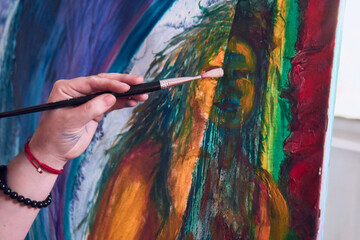 Fototapeta premium View of a woman's hands painting a picture. Next to the window, on a sunny day, multicolored paint, moving the brush, next to several brushes on her clothing, attitude, and beauty.