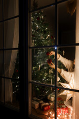 Merry Christmas and Happy holidays! Happy woman in santa hat and cozy sweater decorating stylish christmas tree with ornaments and glowing lights in evening in window. Magical xmas eve