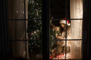 Merry Christmas and Happy holidays! Happy woman in santa hat and cozy sweater decorating stylish christmas tree with ornaments and glowing lights in evening in window. Magical xmas eve