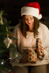 Merry Christmas and Happy holidays! Woman in santa hat and cozy sweater holding shiny gift box at...