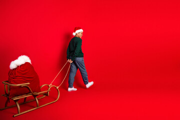 Excited boy dressed as Santa Claus pulls a bag of gifts on a wooden sleigh against a vibrant red background