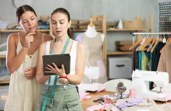Woman fashion designer and a dressmaker discuss sketches of summer dress collections and draw a design on a tablet together. Workers of a sewing workshop draw models of clothes