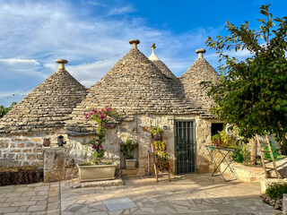 Traditionelles Trulli-Haus in Apulien, Italien, mit typischen Kegeld&auml;chern aus Stein, bl&uuml;henden Pflanzen und mediterranem Ambiente unter blauem Himmel.
