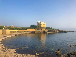 Historisches gelbes K&uuml;stenschloss am Meer bei Alghero auf Sardinien, Italien, umgeben von Felsen und klarem Wasser unter blauem Himmel. Mediterrane Stimmung im Sommer.