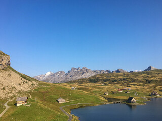 Idyllische Alpenlandschaft mit Bergsee, gr&uuml;nen Wiesen und verstreuten H&uuml;tten unter klarem blauem Himmel &ndash; friedliche Sommerstimmung in den Schweizer Alpen.