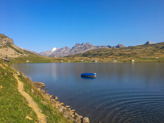 Idyllischer Bergsee mit klarem Wasser, grasbewachsenen H&uuml;geln und schneebedeckten Gipfeln im Hintergrund. Ein kleiner Pfad f&uuml;hrt am Ufer entlang, friedliche Alpenlandschaft bei Sonnenschein.